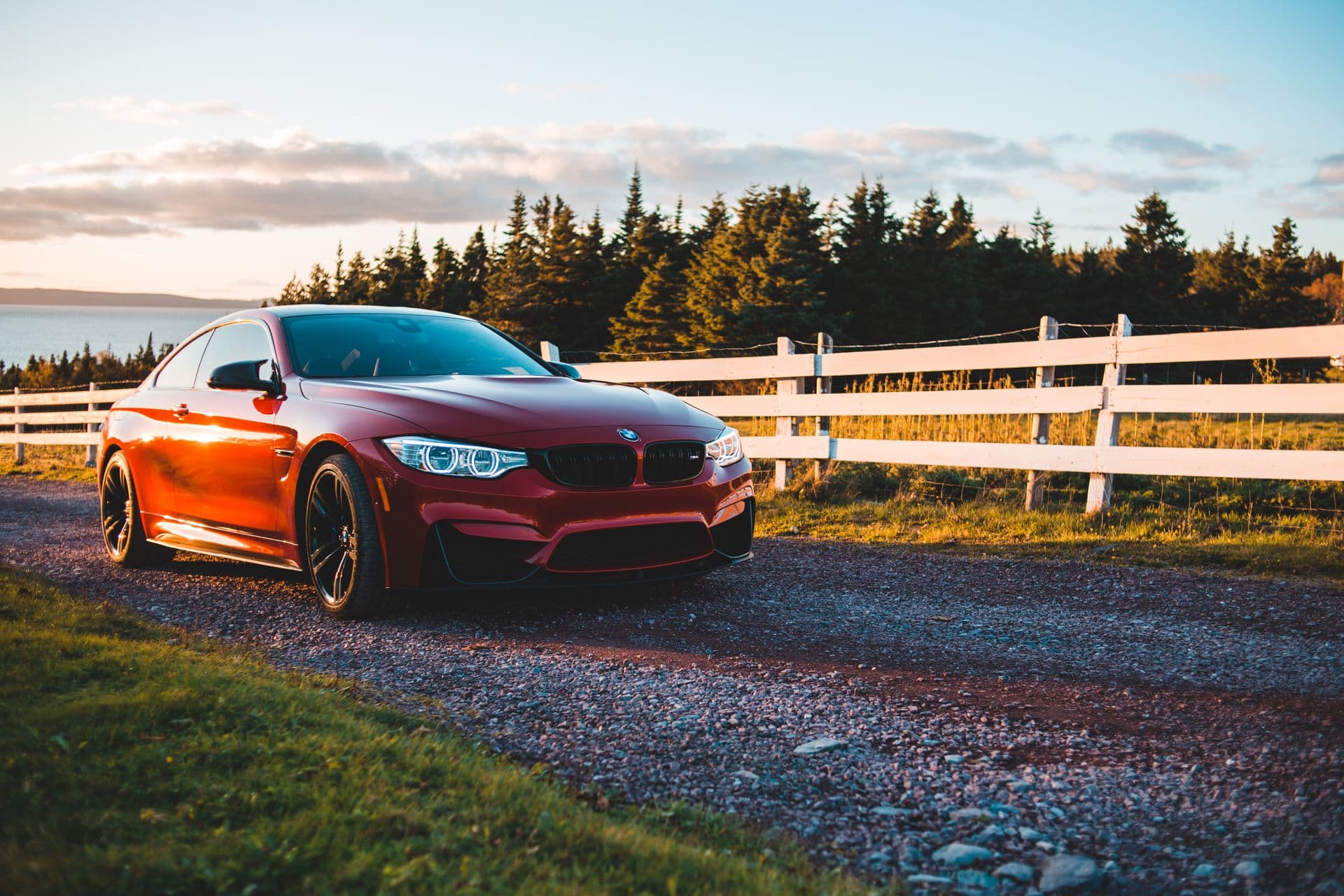 A red coupe parked on a gravel lane by a lake and fir trees at golden hour